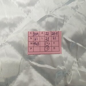 Puede incluir: Una etiqueta rectangular rosa con texto y n&uacute;meros negros se coloca sobre una tela acolchada azul claro. La etiqueta contiene varias filas de n&uacute;meros y letras, incluyendo "369", "22" y "20,5". La tela tiene un patr&oacute;n de diamantes.
