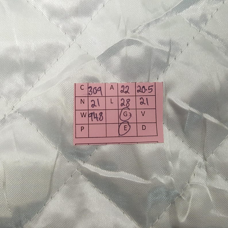 Puede incluir: Una etiqueta rectangular rosa con texto y n&uacute;meros negros se coloca sobre una tela acolchada azul claro. La etiqueta contiene varias filas de n&uacute;meros y letras, incluyendo "369", "22" y "20,5". La tela tiene un patr&oacute;n de diamantes.