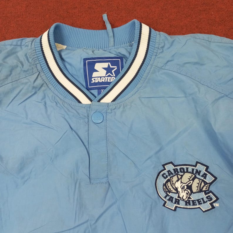 Pu&ograve; includere: Giacca pullover Starter azzurra con colletto a righe bianche e blu navy. La giacca ha una chiusura con bottoni e una toppa con il logo Carolina Tar Heels. Il logo Starter &egrave; visibile sul colletto.