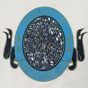 May include: White t-shirt featuring a large, oval emblem with the text "UNIVERSITY OF NORTH CAROLINA" in light blue. The emblem has a floral design in the center, with the letters "UNC" in the middle. Dark blue ribbons are on either side.