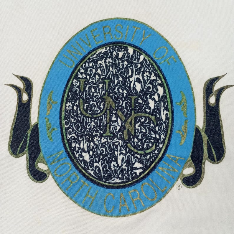 May include: White t-shirt featuring a large, oval emblem with the text "UNIVERSITY OF NORTH CAROLINA" in light blue. The emblem has a floral design in the center, with the letters "UNC" in the middle. Dark blue ribbons are on either side.