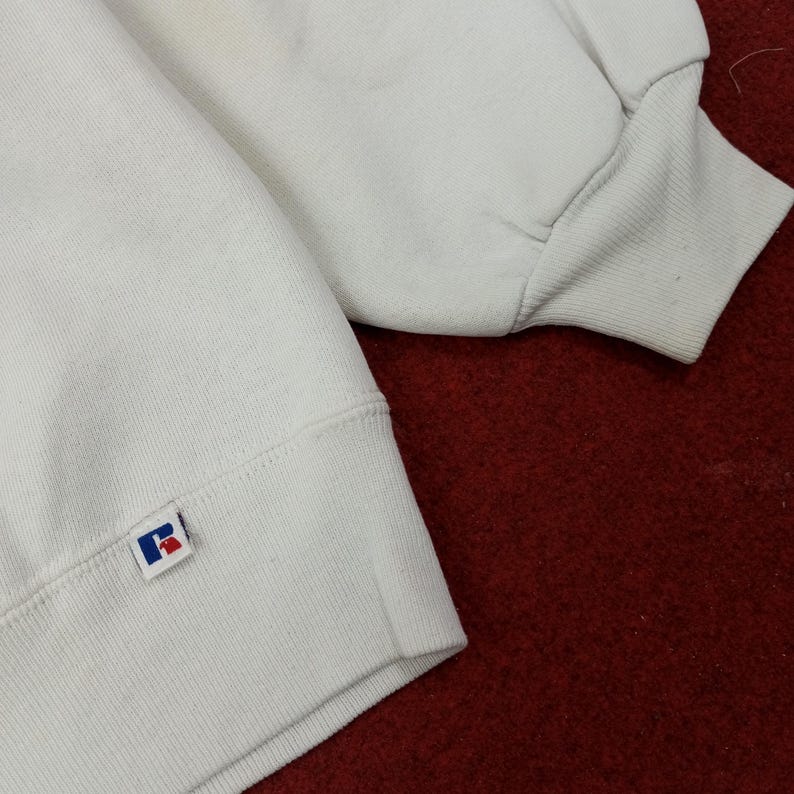 May include: Close-up of a white sweatshirt with ribbed cuffs and a crew neck. The sweatshirt features a small blue and red logo patch on the lower left sleeve. The fabric appears to be a soft, comfortable material.
