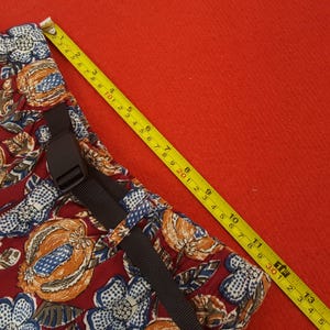 May include: A section of patterned fabric with a black buckle and strap. The fabric is a deep red with a floral design in blue, orange, and white. A yellow measuring tape shows a length of approximately 33 cm.