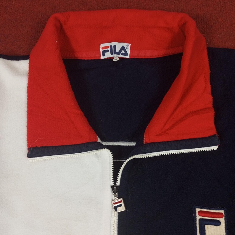 May include: A blue, white, and red zip-up fleece jacket with the FILA logo on the front. The jacket has a high collar and a zipper closure.