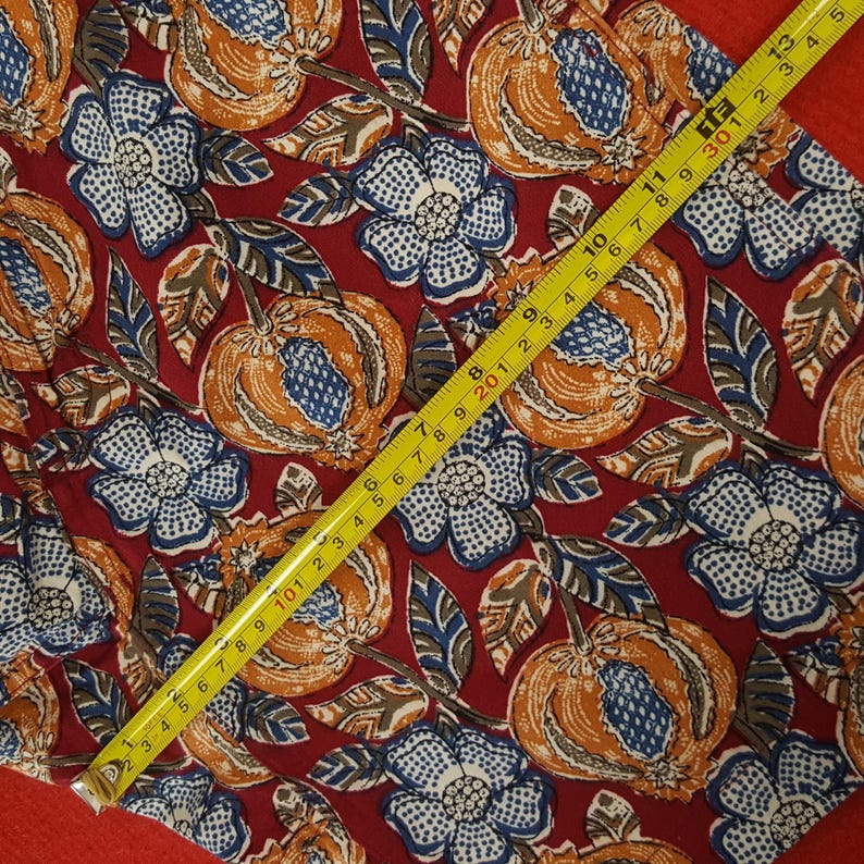 May include: A fabric featuring a repeating pattern of orange fruit and blue and white flowers on a burgundy background. A yellow tape measure shows a measurement of 33 cm.