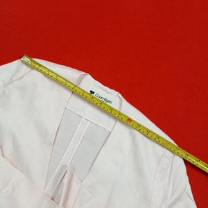 May include: Pale pink Courr&egrave;ges jacket with a V-neck design. A yellow measuring tape is laid across the jacket, showing measurements in inches. The jacket has a clean, minimalist aesthetic, with the Courr&egrave;ges logo visible on the label.