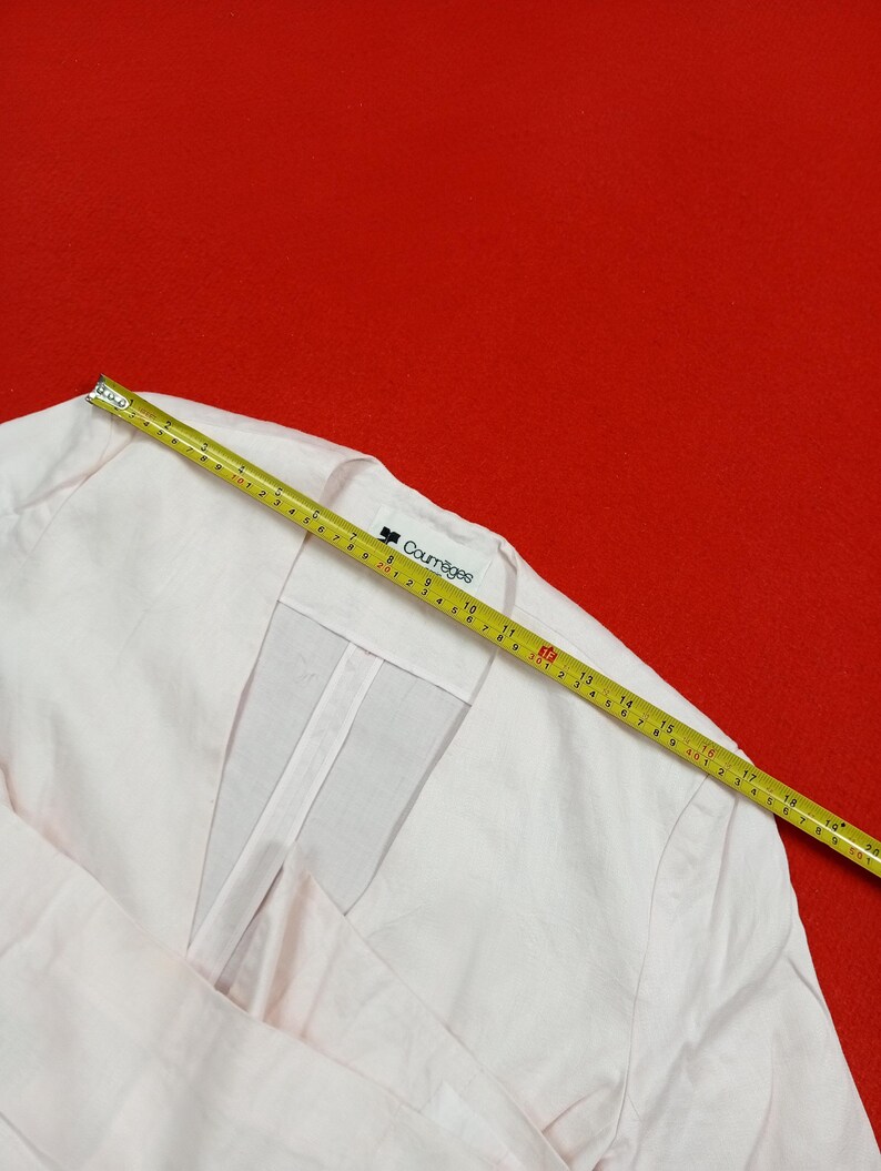 May include: Pale pink Courr&egrave;ges jacket with a V-neck design. A yellow measuring tape is laid across the jacket, showing measurements in inches. The jacket has a clean, minimalist aesthetic, with the Courr&egrave;ges logo visible on the label.