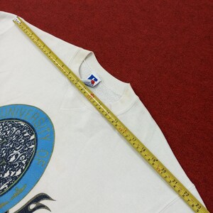 May include: A white crew neck sweatshirt with a blue and black graphic design. The sweatshirt has a vintage look and feel. A measuring tape is laid across the sweatshirt, showing measurements in inches.