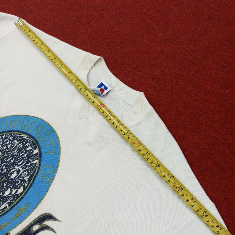 May include: A white crew neck sweatshirt with a blue and black graphic design. The sweatshirt has a vintage look and feel. A measuring tape is laid across the sweatshirt, showing measurements in inches.