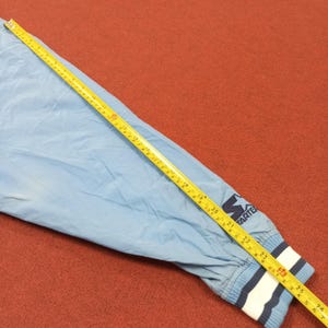 Pu&ograve; includere: Manica di giacca Starter azzurra con polsino a righe bianche, blu e navy. La giacca ha un logo Starter blu navy. Un metro a nastro giallo &egrave; appoggiato sulla manica, con misure in centimetri.