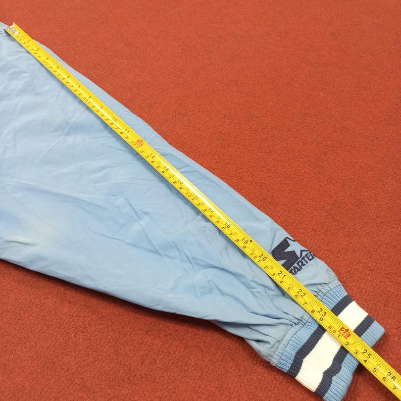 Pu&ograve; includere: Manica di giacca Starter azzurra con polsino a righe bianche, blu e navy. La giacca ha un logo Starter blu navy. Un metro a nastro giallo &egrave; appoggiato sulla manica, con misure in centimetri.