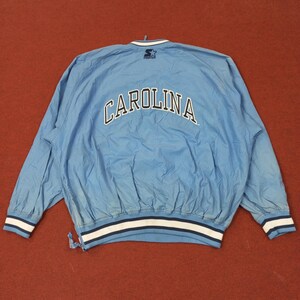 Pu&ograve; includere: Giacca a vento azzurra con la scritta "CAROLINA" ad arco sulla schiena in lettere bianche. La giacca ha un bordo a righe bianche e blu navy sui polsini e in vita. Il logo Starter &egrave; visibile sul colletto.