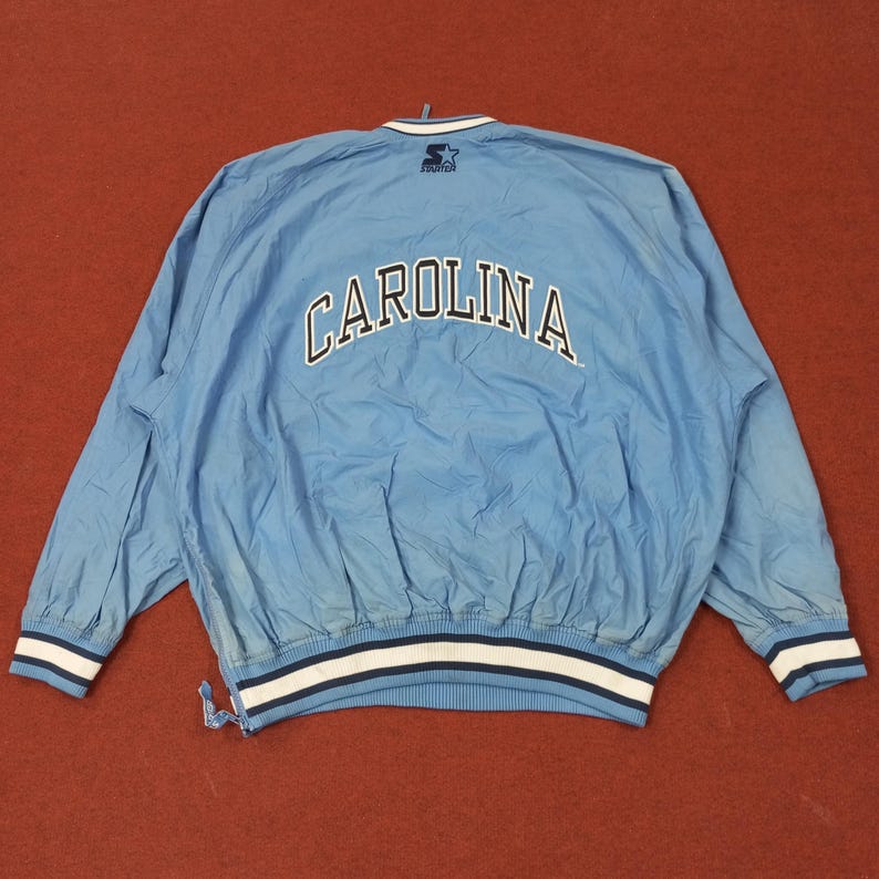 Pu&ograve; includere: Giacca a vento azzurra con la scritta "CAROLINA" ad arco sulla schiena in lettere bianche. La giacca ha un bordo a righe bianche e blu navy sui polsini e in vita. Il logo Starter &egrave; visibile sul colletto.