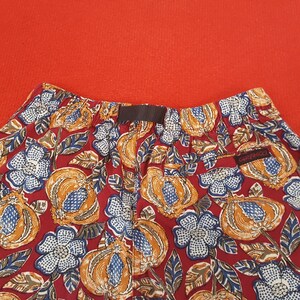 May include: Close-up of patterned shorts against a red background. The shorts display a repeating pattern of orange and blue fruit and white and blue flowers on a red base. The shorts have a brown waistband and a small "GRAMICCI" tag.