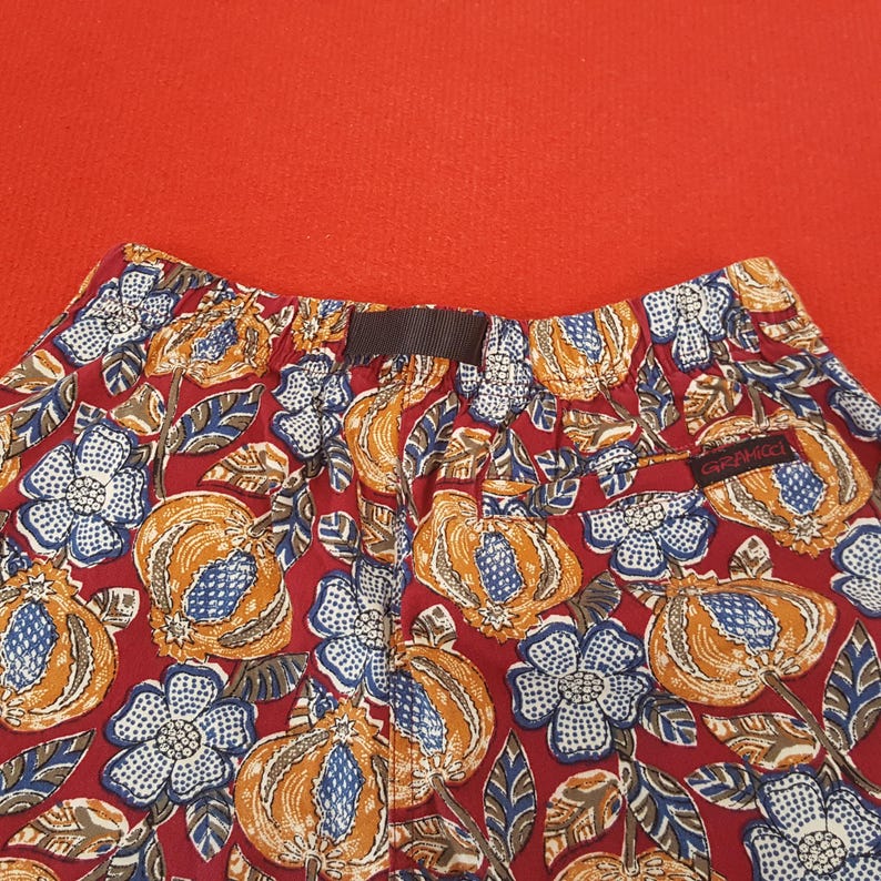 May include: Close-up of patterned shorts against a red background. The shorts display a repeating pattern of orange and blue fruit and white and blue flowers on a red base. The shorts have a brown waistband and a small "GRAMICCI" tag.