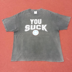 Puede incluir: Una camiseta gris oscuro con las palabras "YOU SUCK" en blanco, delineadas en azul. Un gráfico de estrella azul y blanco está centrado debajo del texto. La camiseta está colocada sobre una superficie roja.