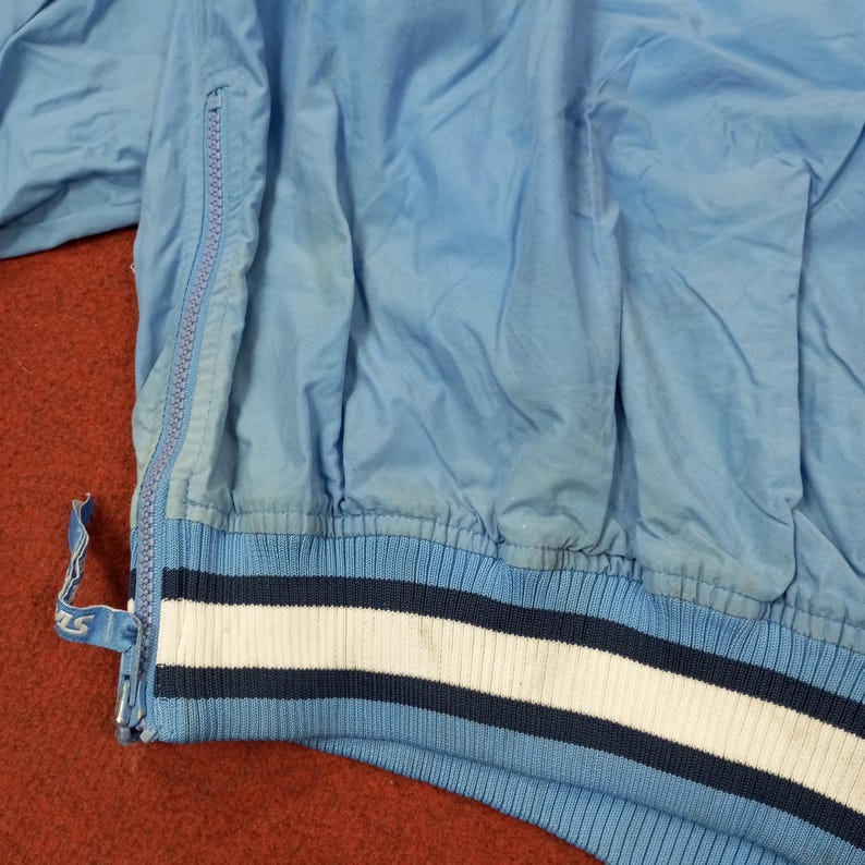 Pu&ograve; includere: Giacca vintage azzurra con chiusura a zip. La giacca presenta orlo e polsini a coste con strisce bianche e blu navy. Il tessuto sembra essere un materiale leggero, forse nylon o poliestere.