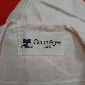 May include: Close-up of a light pink garment with a label that reads "Courr&egrave;ges paris" in black text. The fabric appears to be a soft, possibly linen or cotton blend. The garment has a collar and a folded design.