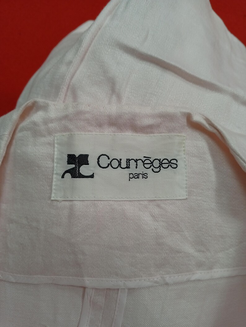 May include: Close-up of a light pink garment with a label that reads "Courr&egrave;ges paris" in black text. The fabric appears to be a soft, possibly linen or cotton blend. The garment has a collar and a folded design.