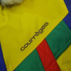Puede incluir: Primer plano de una chaqueta colorida con un panel amarillo con el nombre de la marca "Courr&egrave;ges" bordado en plata. La chaqueta tiene secciones verdes, rojas y azules. Un ribete blanco y esponjoso es visible en la parte superior. La chaqueta es probablemente vintage.