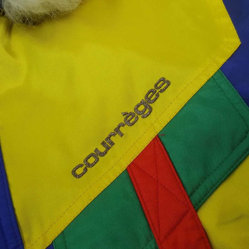Puede incluir: Primer plano de una chaqueta colorida con un panel amarillo con el nombre de la marca "Courr&egrave;ges" bordado en plata. La chaqueta tiene secciones verdes, rojas y azules. Un ribete blanco y esponjoso es visible en la parte superior. La chaqueta es probablemente vintage.
