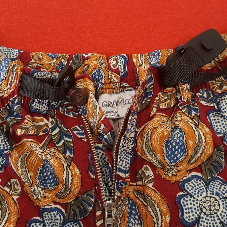 May include: Close-up of patterned trousers with a red background. The trousers feature a floral design in shades of orange, blue, and white on a burgundy base. The brand tag reads "Gramicci Original Freedom". The trousers have a zipper and adjustable straps.