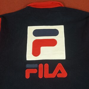May include: A black and red Fila logo on a navy blue jacket. The logo is a white square with a red and blue "F" inside. The word "FILA" is written in red below the square.
