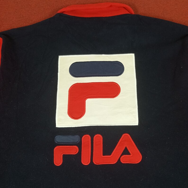 May include: A black and red Fila logo on a navy blue jacket. The logo is a white square with a red and blue "F" inside. The word "FILA" is written in red below the square.