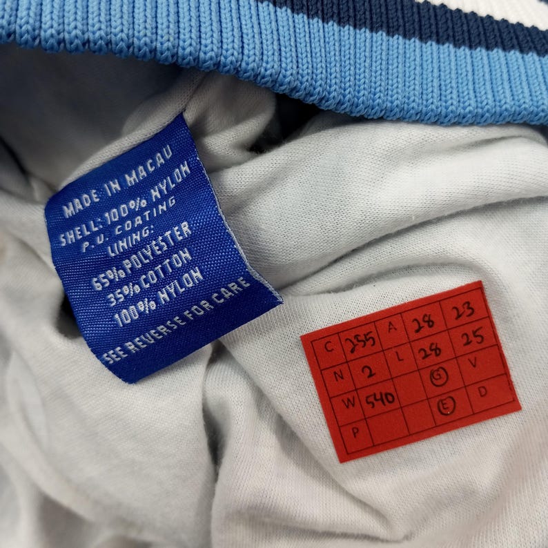 Pu&ograve; includere: Primo piano di un'etichetta blu con testo bianco che recita "MADE IN MACAU" e i dettagli della composizione del capo. &Egrave; visibile anche un'etichetta rossa con numeri e lettere scritte a mano.