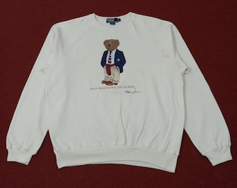 May include: White crew neck sweatshirt featuring a Polo Bear graphic. The bear is dressed in a navy blazer, red tie, and khaki pants. The text "POLO BEAR by RALPH LAUREN" is printed below the bear, with a signature. A small logo is visible on the upper back.