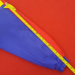 Puede incluir: Una chaqueta colorida en azul, rojo y amarillo, con una cinta m&eacute;trica amarilla colocada encima. La manga de la chaqueta est&aacute; fruncida en la mu&ntilde;eca. La cinta m&eacute;trica muestra medidas de hasta 56 cm.