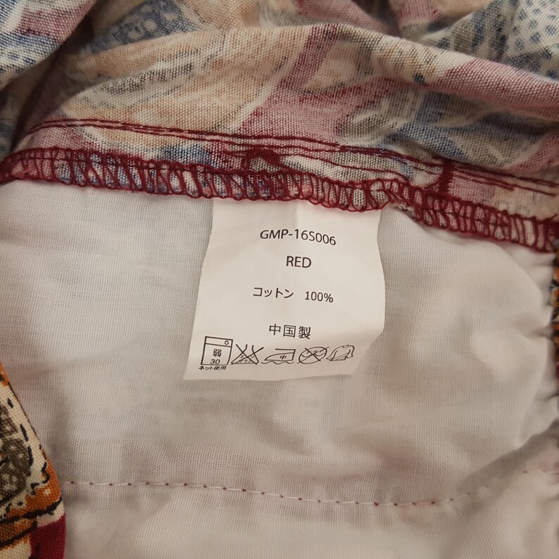 May include: A close-up of a garment label with the text "GMP-165006 RED コットン 100% 中国製". The label is white with black text and is attached to a red and patterned fabric. The fabric has a mix of red, blue, and tan colours.