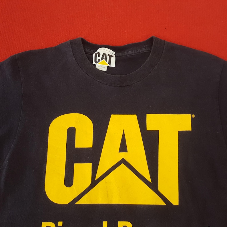 May include: A navy blue t-shirt with a large yellow "CAT" logo and a smaller logo on the collar. The shirt has a crew neck and is made of a soft material. The background is a solid red.