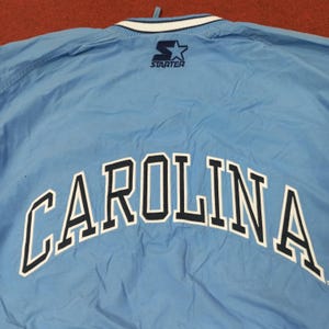 Pu&ograve; includere: Giacca Starter azzurra con la scritta "CAROLINA" ad arco sulla schiena in bianco con bordi blu navy. Il logo Starter &egrave; visibile sulla parte superiore della schiena. Il colletto ha strisce bianche e blu navy.