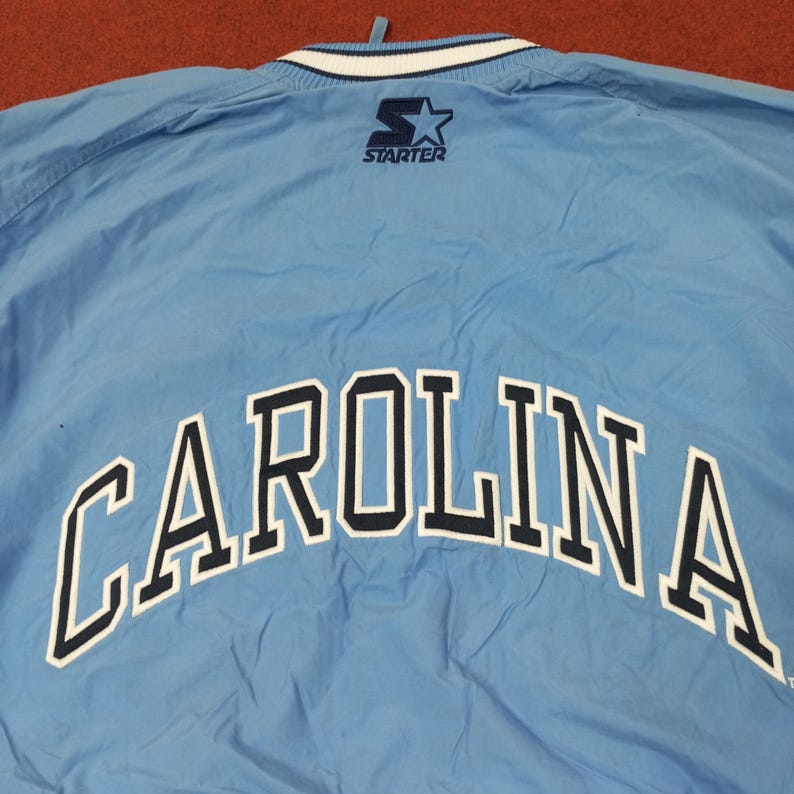 Pu&ograve; includere: Giacca Starter azzurra con la scritta "CAROLINA" ad arco sulla schiena in bianco con bordi blu navy. Il logo Starter &egrave; visibile sulla parte superiore della schiena. Il colletto ha strisce bianche e blu navy.