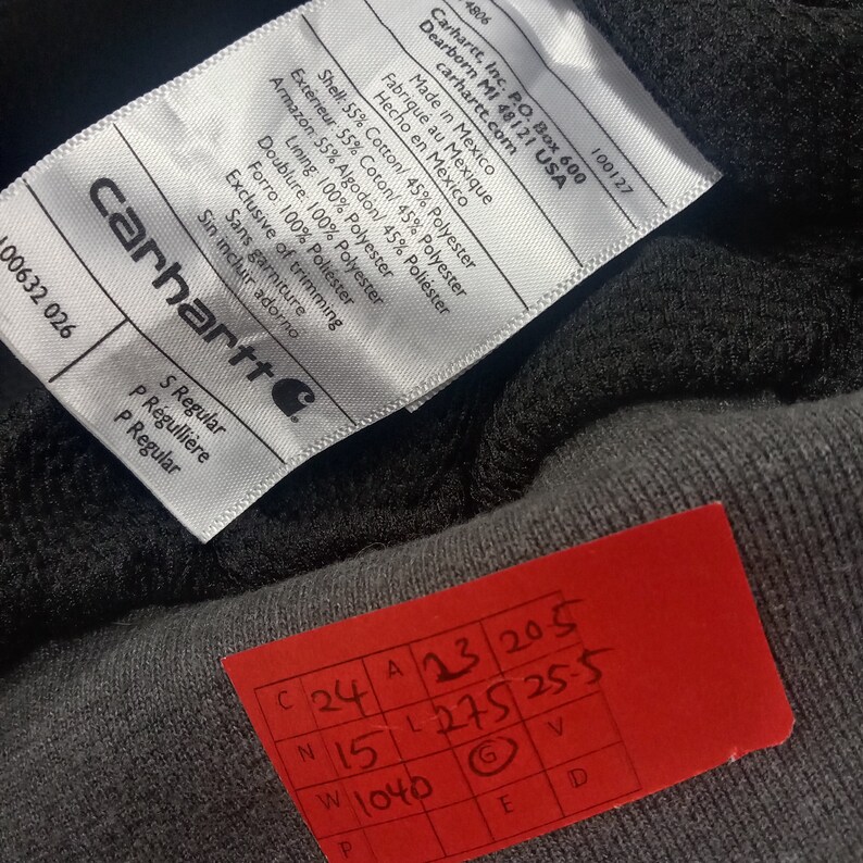May include: A white tag with black text attached to a black and gray garment. The tag lists the garment's material composition, country of origin, and size. The tag also includes the Carhartt logo and the text "S Regular, P R&eacute;gulli&egrave;re, P Regular". A red tag with black text is also visible, with the text "C 24 A 13 20 5 N 15 L 275 25 5 W 1040 P G V E D".