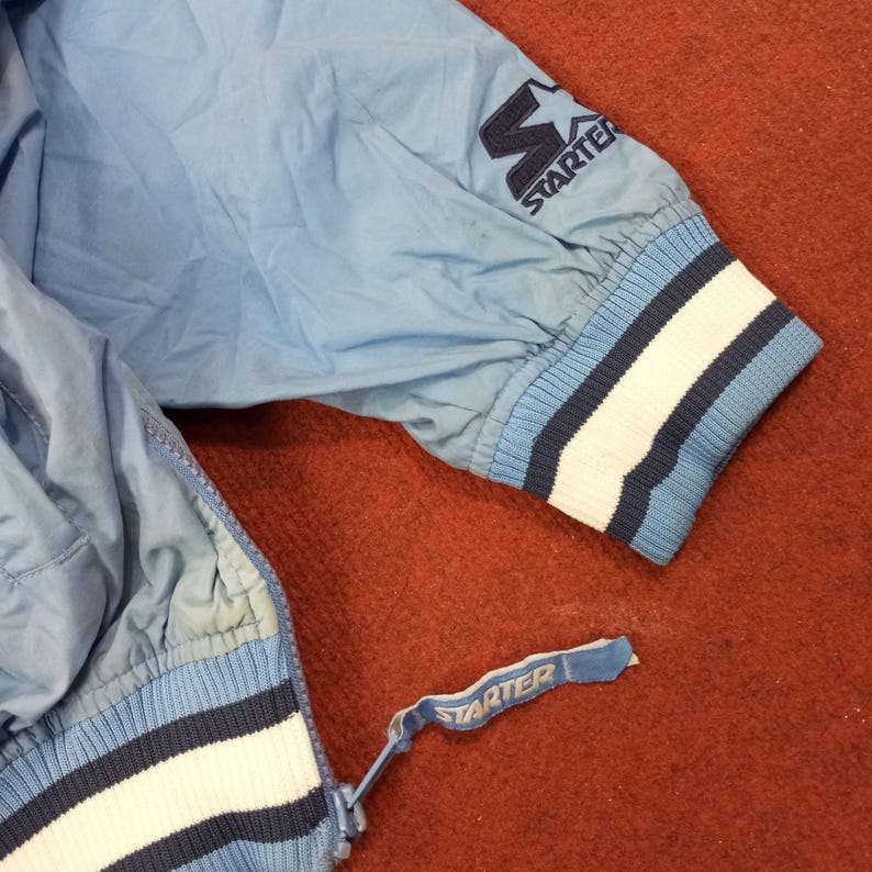 Pu&ograve; includere: Giacca Starter azzurra con cerniera e polsino a coste. Il polsino ha strisce bianche e blu navy. Il logo Starter &egrave; sulla manica e sul tirazip.