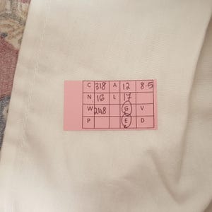 May include: A pink tag with handwritten numbers and letters is placed on a white fabric. The tag includes the following information: C318, A12, 8-5, N16, L17, W248, P, G, E, V, D. The fabric is white.