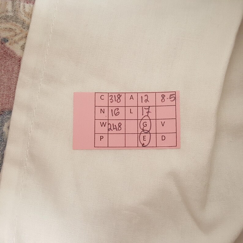 May include: A pink tag with handwritten numbers and letters is placed on a white fabric. The tag includes the following information: C318, A12, 8-5, N16, L17, W248, P, G, E, V, D. The fabric is white.