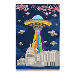 May include: A dark blue towel featuring a whimsical illustration of a UFO beaming up cows over the U.S. Capitol building. The design includes a rainbow beam, flying saucers, cherry blossoms, and stars. The towel is a fun and unique home decor item.