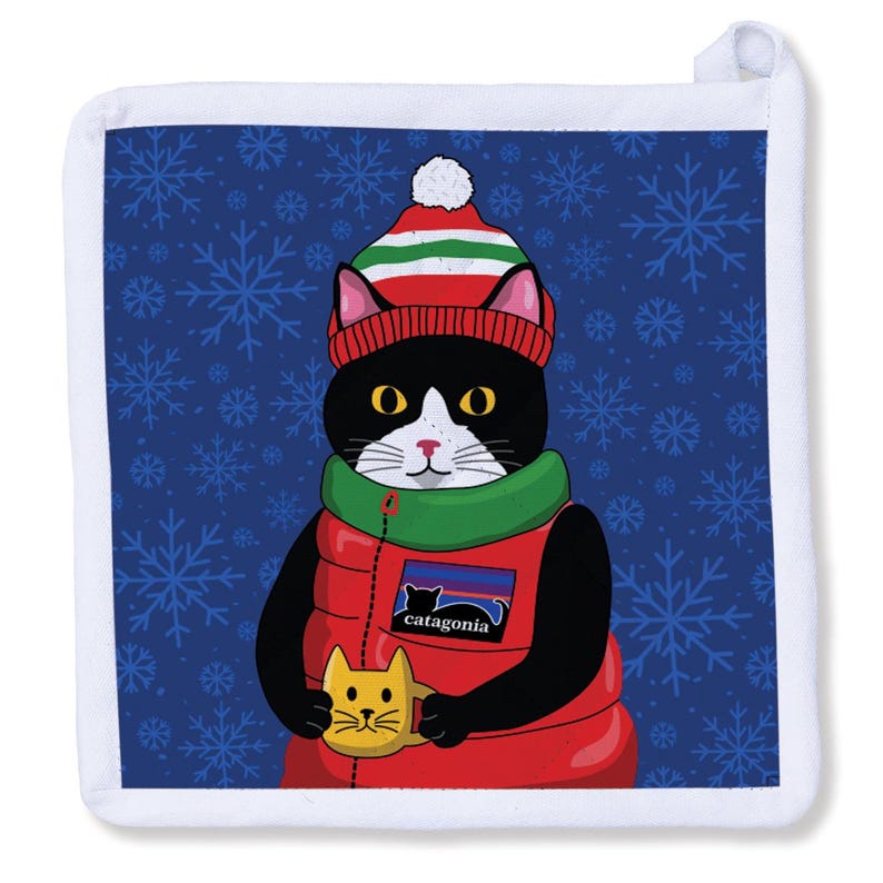 May include: A square pot holder with a white border. It depicts a black and white cat in a red puffer vest, green scarf, and a striped hat. The cat holds a yellow mug with a cat face. The background is blue with white snowflakes, and the vest has the word "catagonia".