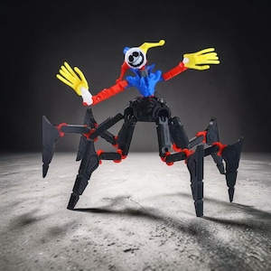 Pode incluir: Uma figura de brinquedo articulada e colorida que se assemelha a um híbrido de palhaço e aranha. Apresenta uma cabeça com rosto branco e expressão sorridente, uma gola azul com babados, braços vermelhos, mãos amarelas e pernas pretas com articulações vermelhas. A figura está sobre uma superfície cinza.