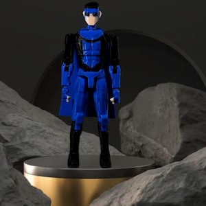 Op de afbeelding: Een blauw actiefiguur met een zwarte cape en laarzen staat op een goud en grijs platform. Het figuur heeft een geometrisch ontwerp met een wit gezicht en zwart haar. De achtergrond bevat grijze rotsen en een donkergrijze boog.