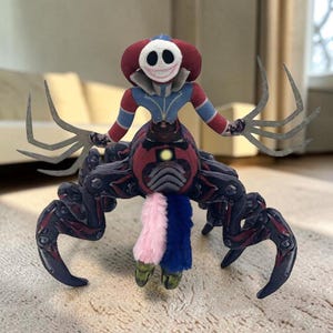 Puede incluir: Un peluche que representa una criatura sonriente, vestida de rojo y azul. Tiene múltiples brazos y un cuerpo rojo oscuro con patas negras y rojas. El juguete tiene una cola rosa, azul y verde.