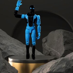 Op de afbeelding: Een blauw en zwart beweegbaar actiefiguur van een humano&iuml;de robot, met &eacute;&eacute;n arm omhoog. Het figuur staat op een goudkleurig platform, tegen een donkere achtergrond met rotsformaties.