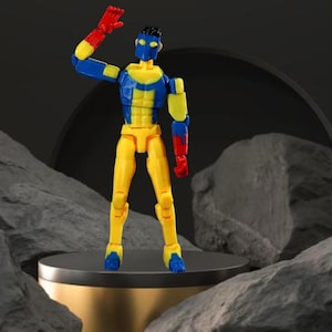 Op de afbeelding: Een kleurrijk actiefiguur met een gele romp en benen, blauwe en gele armen en rode handen. Het figuur heeft een blauw hoofd en poseert met &eacute;&eacute;n arm omhoog. Het staat op een klein, rond platform.