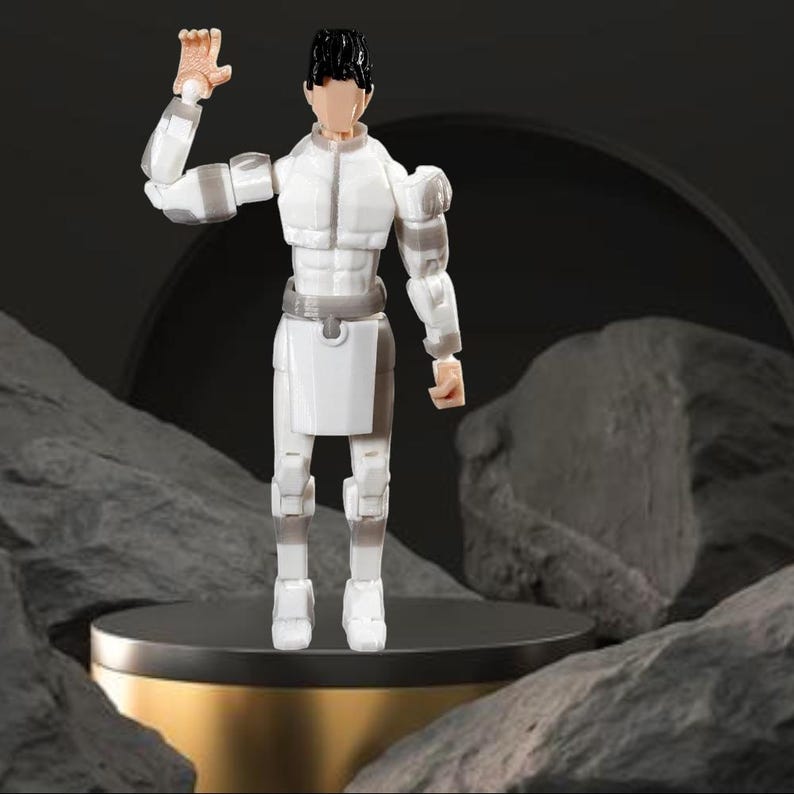 Op de afbeelding: Een wit actiefiguur van een persoon in een futuristisch kostuum, staand op een goud en zwart platform. Het figuur heeft zwart haar, een bruin gezicht en heft zijn rechterhand op. Het figuur is omgeven door grijze rotsformaties.