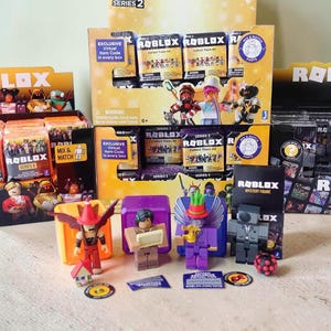 May include: A display of Roblox Series 2 collectible figures and blind boxes. The figures are brightly colored and include characters with unique designs. The packaging features the Roblox logo and the words "Mix & Match Parts."