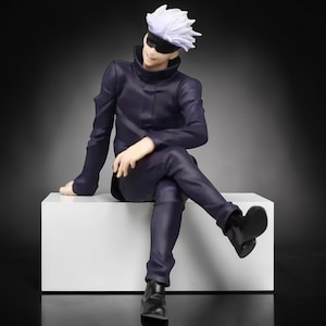 May include: A figurine of a character with white spiky hair, a black blindfold, and a dark purple outfit, seated on a white block. The figure is posed with one leg crossed over the other, and black boots.