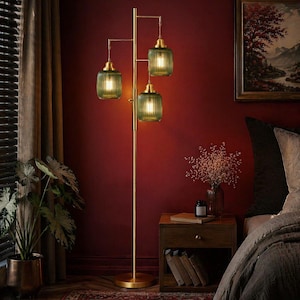 May include: A gold-toned floor lamp with three green glass shades. The lamp has a tall, slender frame with a round base. The shades are rectangular with vertical ridges. The lamp is in a room with a dark red wall.
